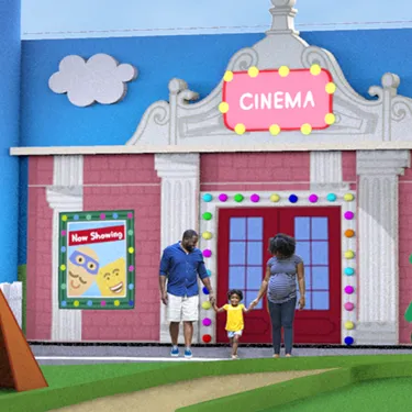 A mom and dad hold hands with their child in front of the Cinema at Peppa Pig Theme Park in Dallas Fort Worth. The family smiles as they stand together, enjoying the vibrant atmosphere of the park with the colorful cinema sign in the background.