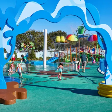 Children and parents enjoy the Muddy Puddles Splash Pad at Peppa Pig Theme Park in Dallas Fort Worth. The kids splash around in the water while the parents watch and laugh, all surrounded by the playful, water-filled atmosphere of the park.