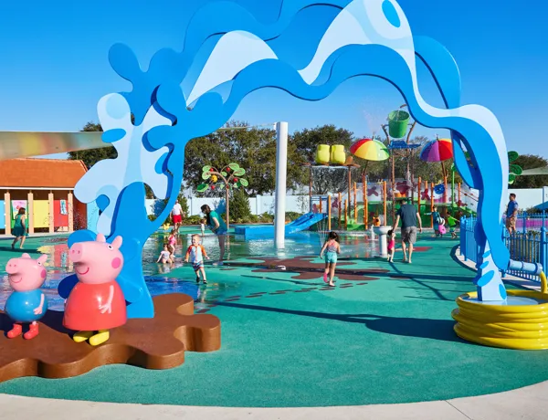 Children and parents enjoy the Muddy Puddles Splash Pad at Peppa Pig Theme Park in Dallas Fort Worth. The kids splash around in the water while the parents watch and laugh, all surrounded by the playful, water-filled atmosphere of the park.