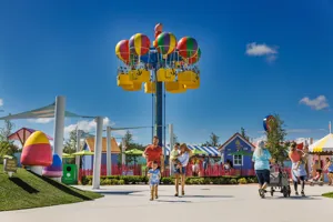 A group of families enjoys the Fun Fair attraction at Peppa Pig Theme Park in Dallas Fort Worth, playing games and riding various attractions. In the background, Peppa's Balloon ride can be seen, adding to the vibrant and playful atmosphere of the park.